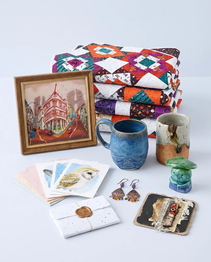 Handmade work on a pale tabletop: folded quilts, a framed painting, a hand-thrown mug and cup, a ceramic figure, notecards, earrings and a collage