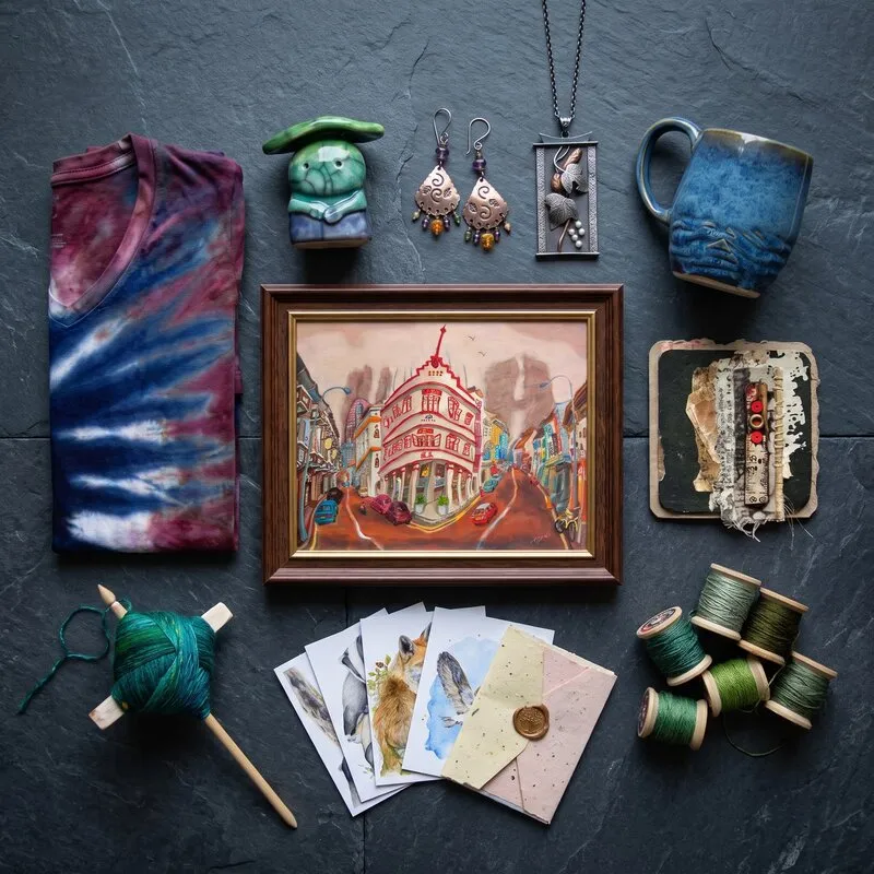 Many kinds of handmade work on dark slate: a tie-dyed shirt, a framed painting, a mug, a ceramic figure, earrings, a pendant, a collage, notecards, yarn and thread