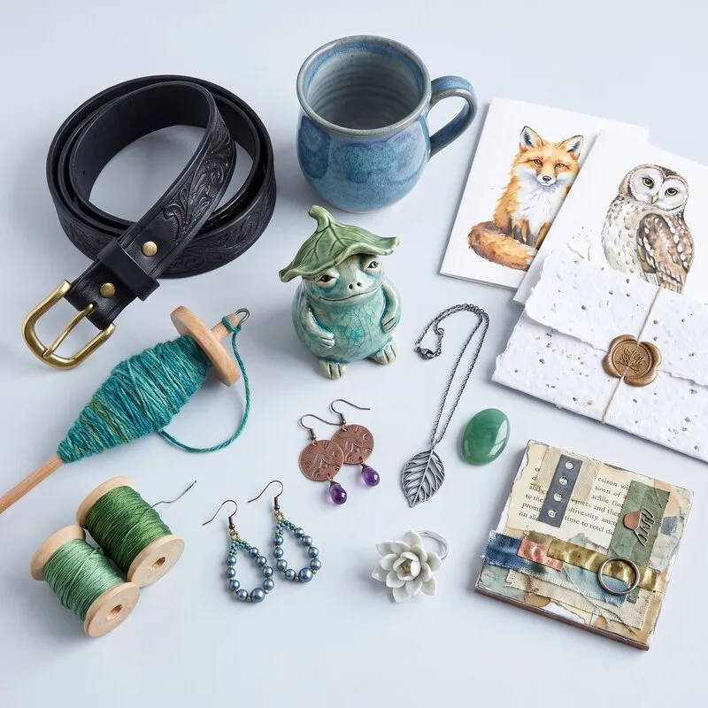 A flat lay of handmade work: a tooled belt, a hand-thrown mug, a ceramic frog, notecards, a spindle of yarn, thread spools, earrings and a collage
